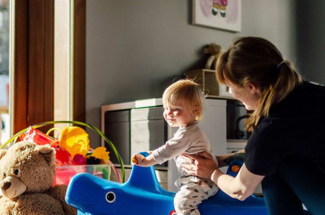 Smiling baby on rocking toy with nanny in Bear Cubs playroom