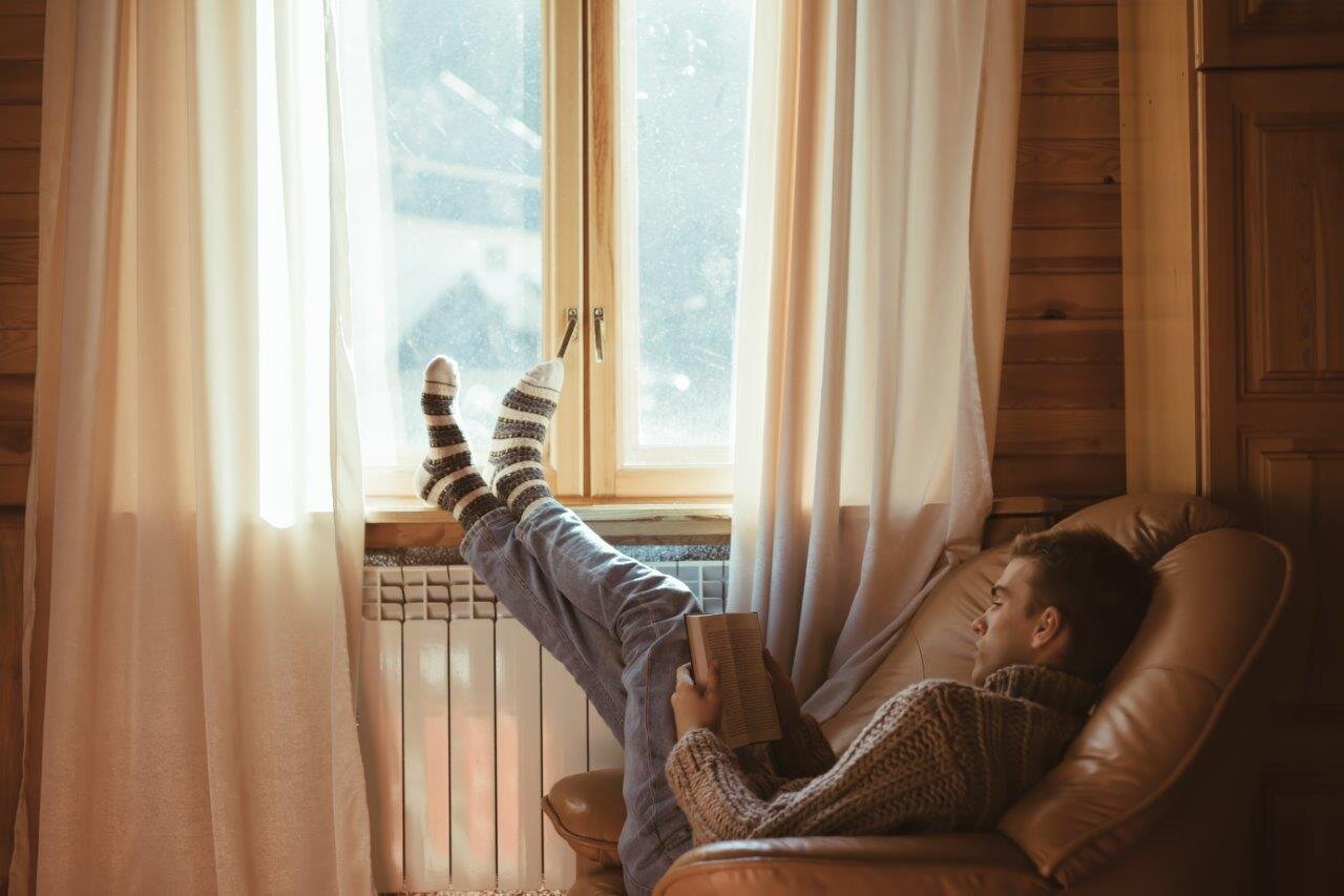 Young man reading book in cosy leather armchair by window with socked feet resting on window sill