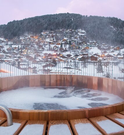 Outdoor wooden hot tub in the evening with the lights of Morzine chalets in the background