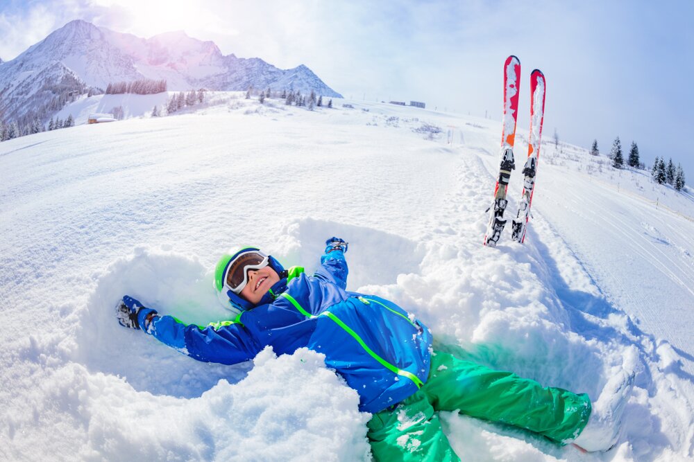 Young boy in ski gear doing snow angel on side of piste