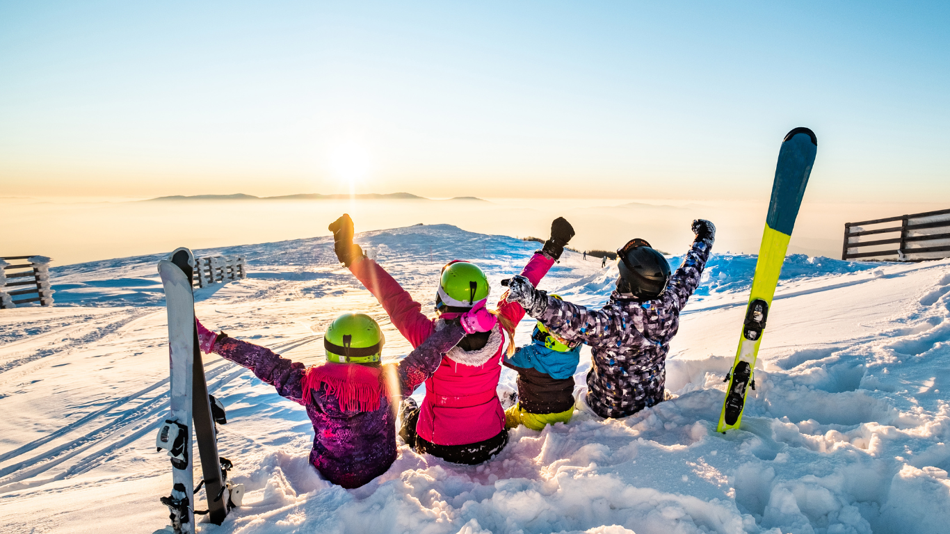 Family of four sitting in snow next to skis with hands in the air looking at the sun