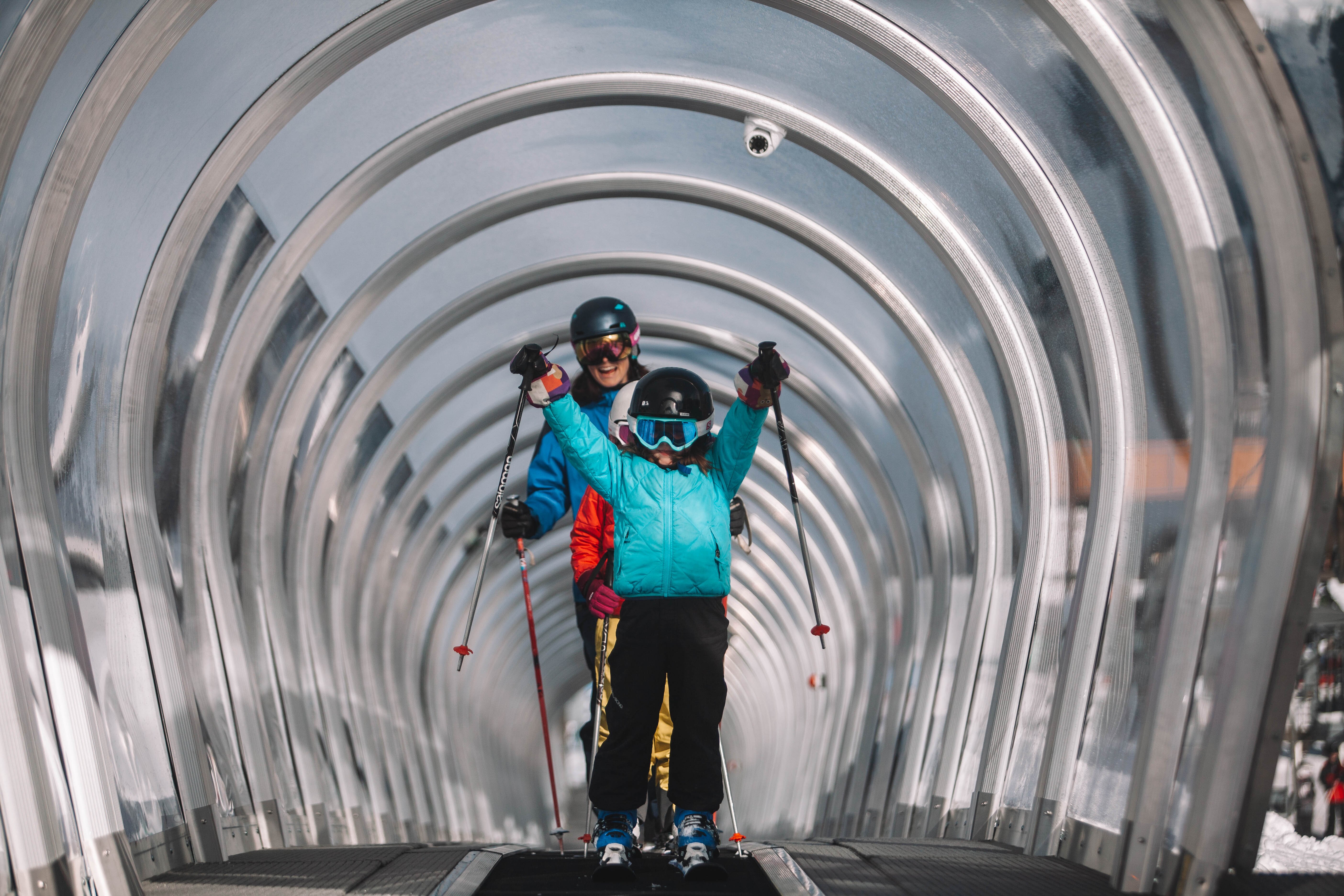 Child holding ski poles in the air whilst coming up covered magic carpet ski lift