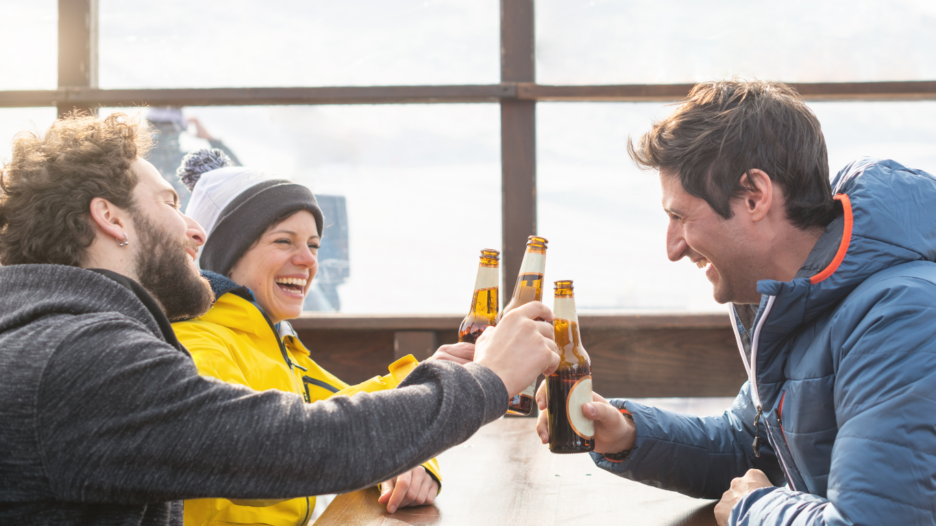 3 smiling friends chinking beer bottles on terrace of outdoor ski bar
