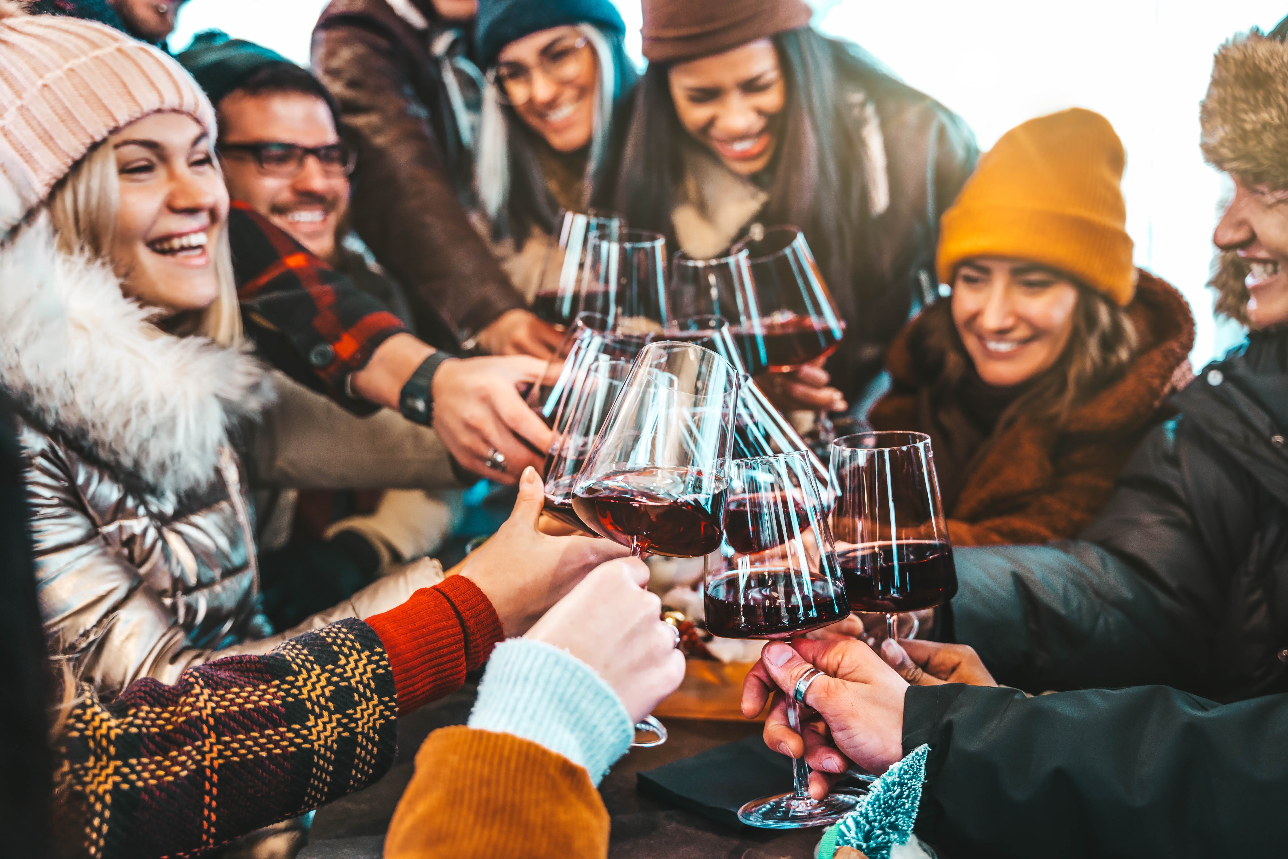Group of friends dressed in warm jackets and hats saying cheers with glasses of red wine in outdoor bar