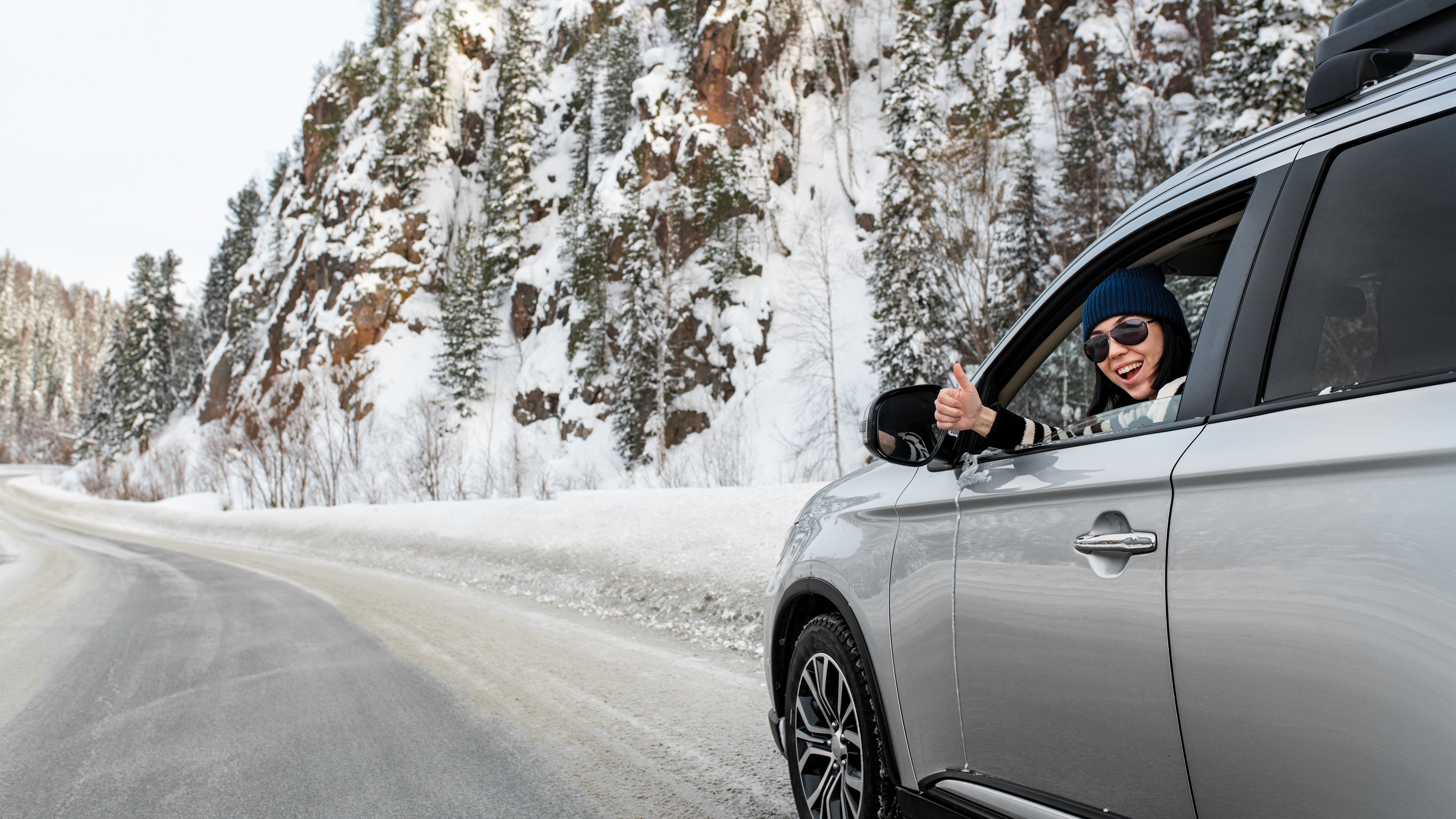 Smiling woman in silver car giving thumbs up through the window on a winding snowy mountain road
