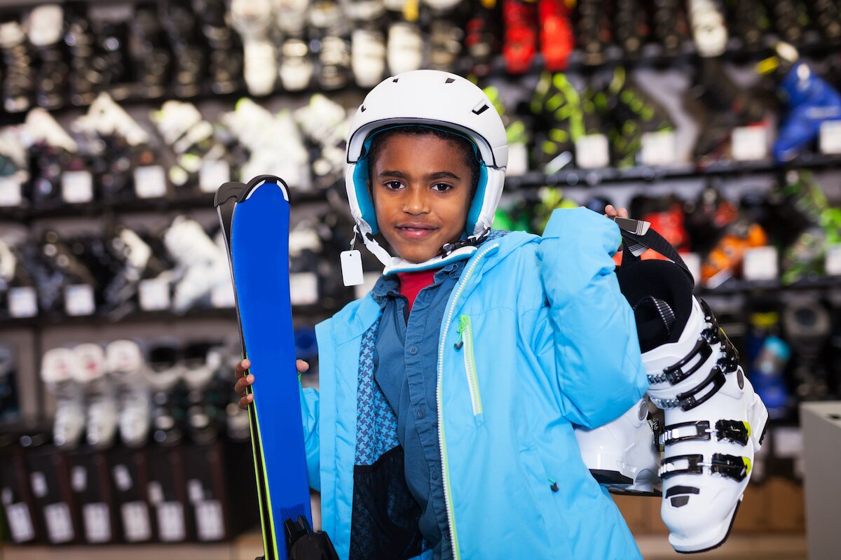 Young boy wearing ski jacket and helmet, holding skis and boots