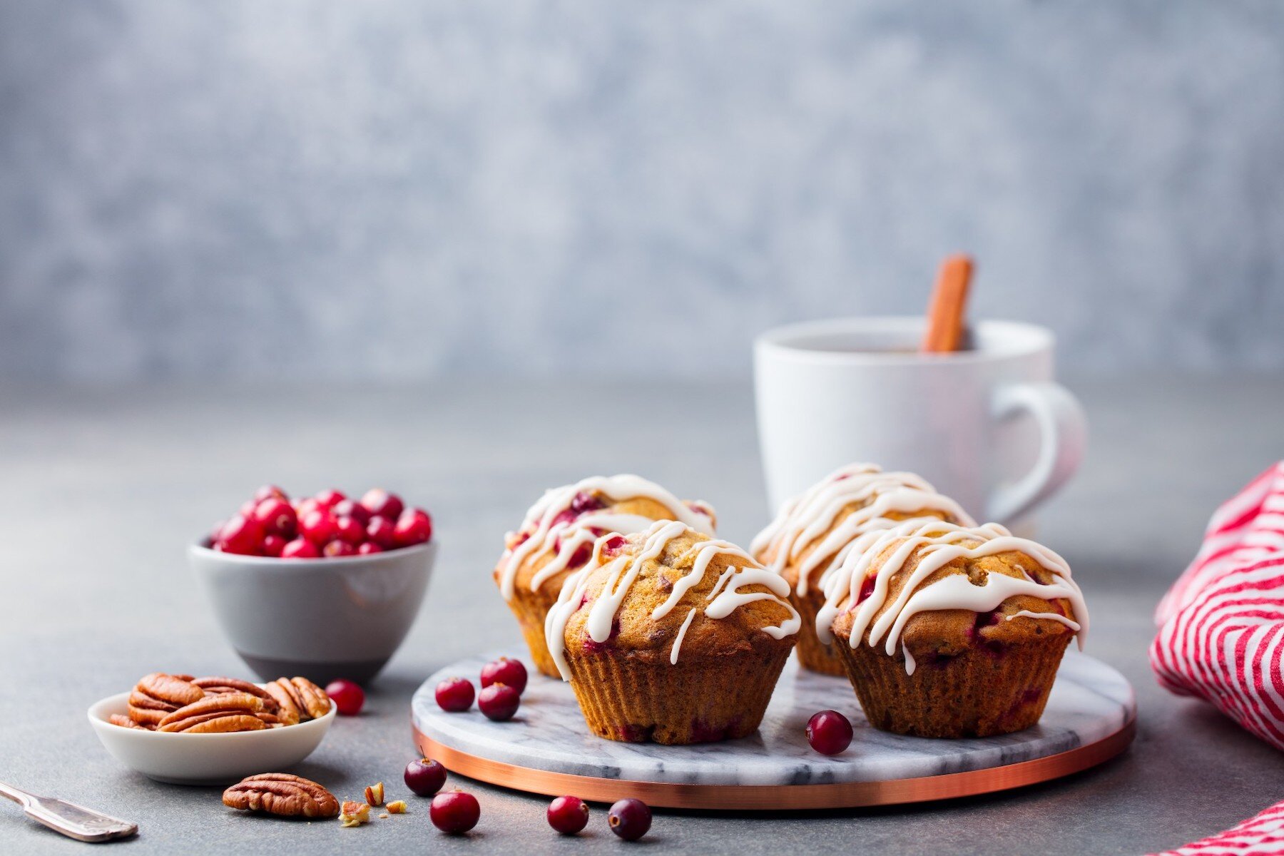 Four iced muffins, pecan nuts, cranberries and a hot chocolate in background