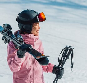 Smiling female skier in pink ski jacket and grey helmet carrying skis and poles