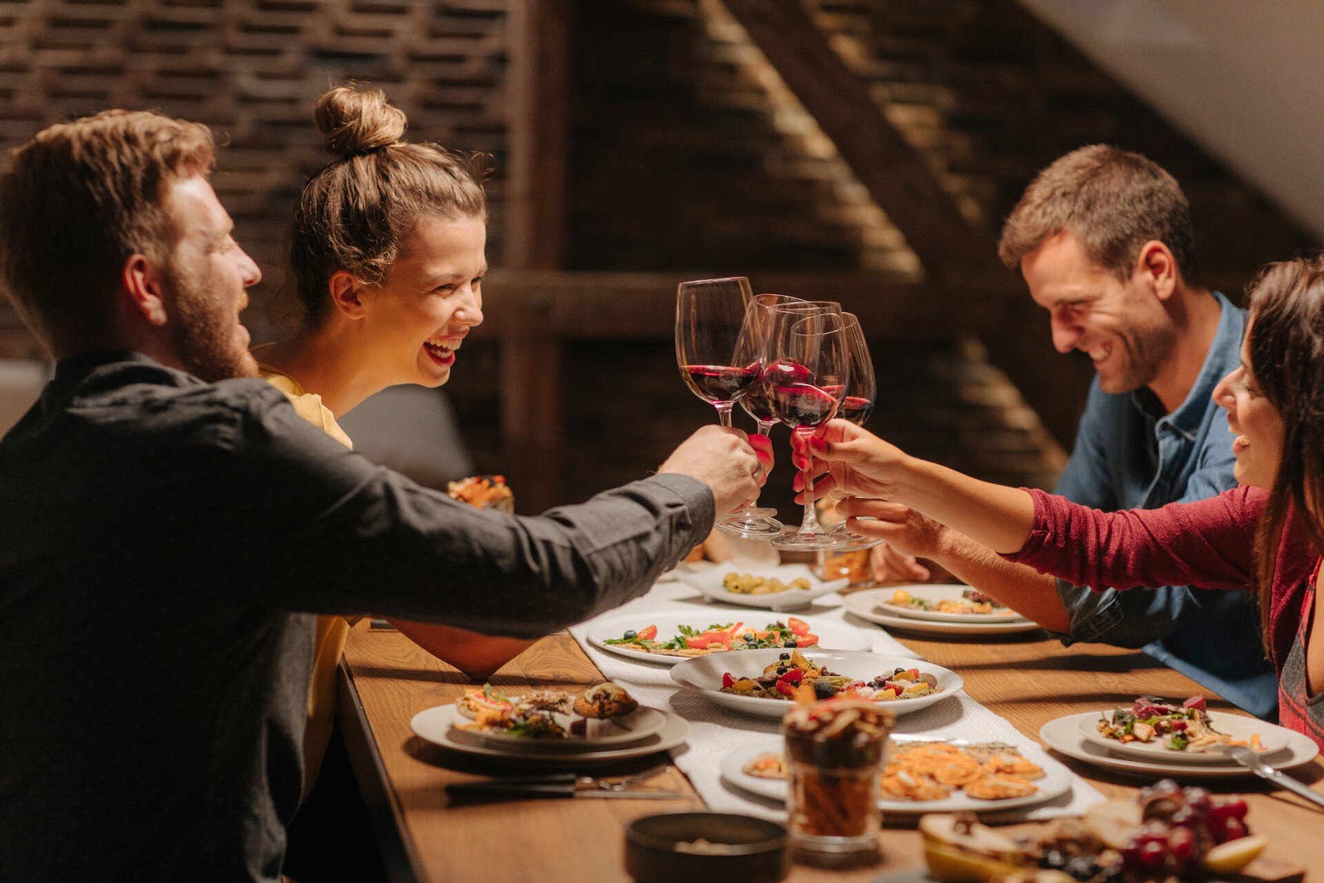 Two smiling couples clinking glasses of red wine together over table of food