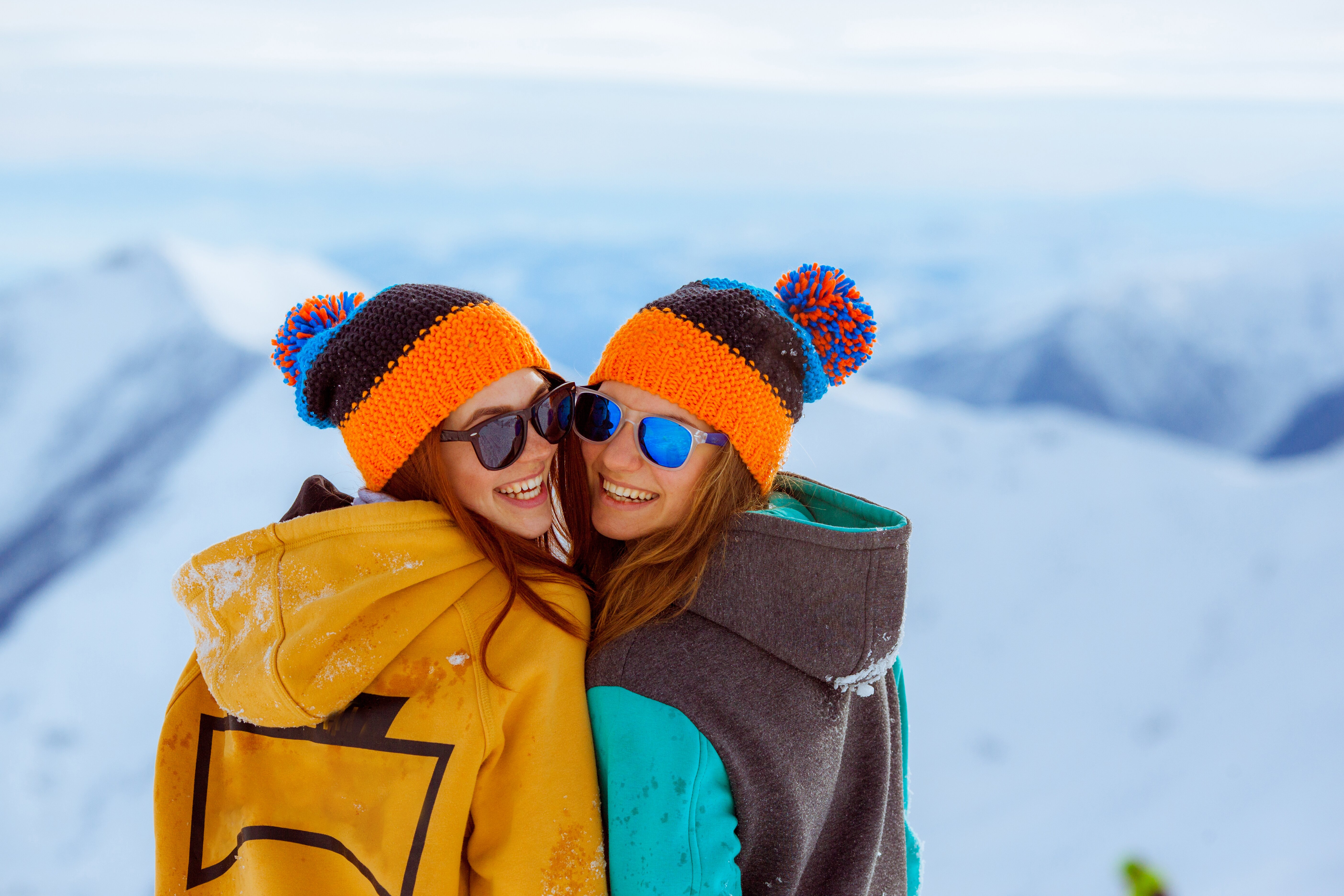 Two smiling women wearing matching bobble hats and sunglasses on snowy slope