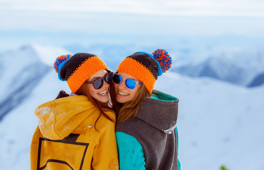 Two smiling women wearing matching bobble hats and sunglasses on snowy slope