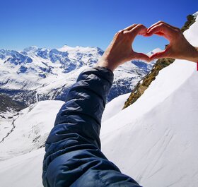 Person making heart shape with hands at the top of the mountain looking down the valley on a sunny day