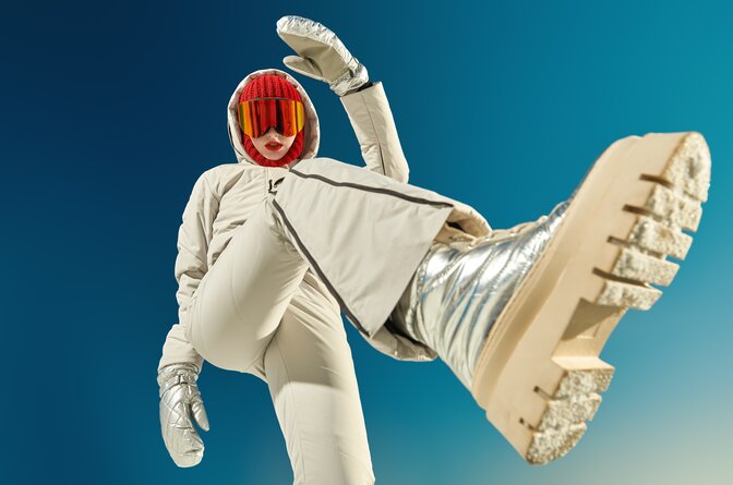 Woman standing over camera wearing beige ski outfit, gold snow boots and a red balaclava