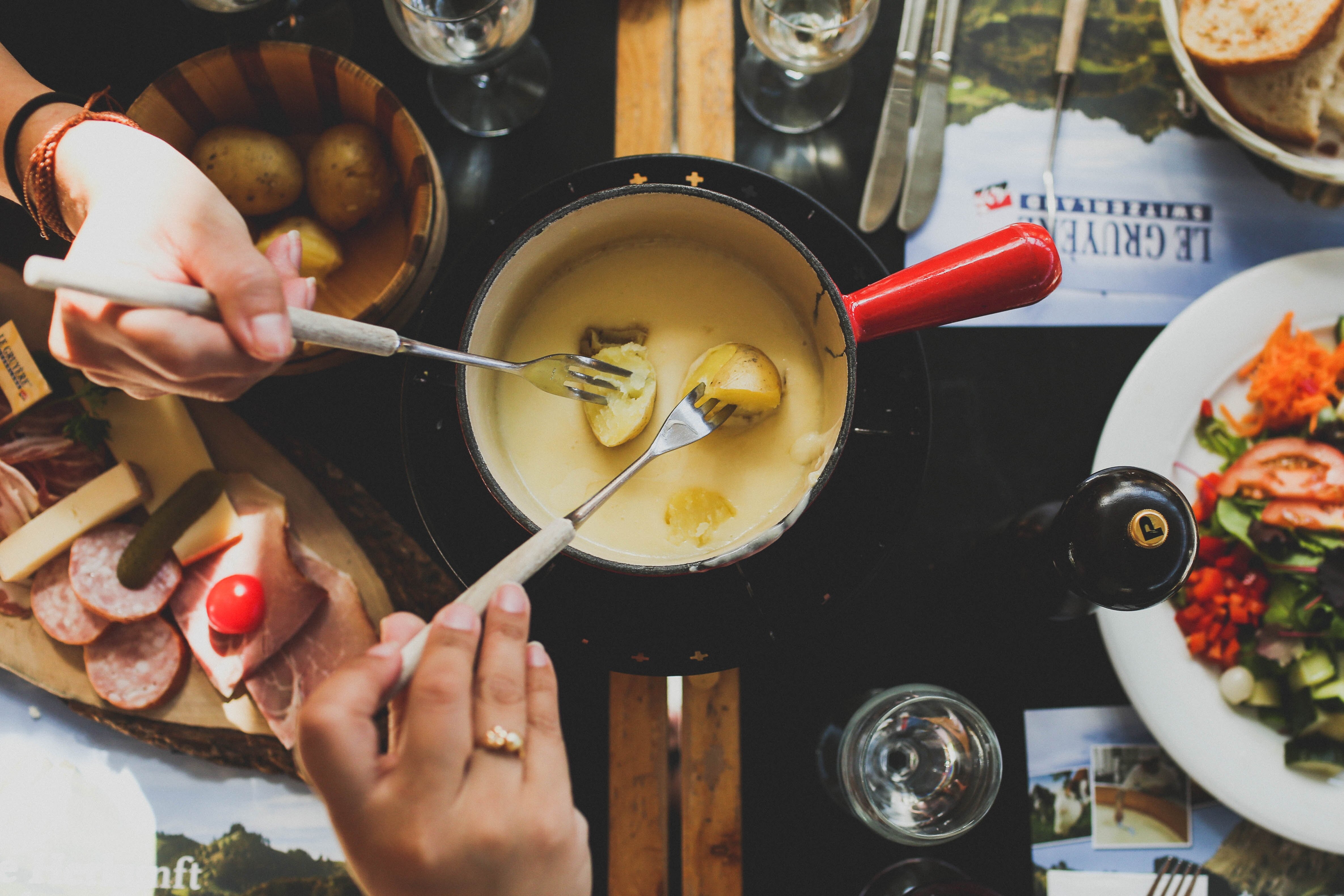 New potatoes being dipped into a cheese fondue with charcuterie and salad on the table