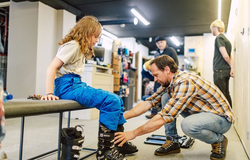 Man in checked shirt fitting ski rental boots to a young girl in the ski room at Bear Lodge ski hotel